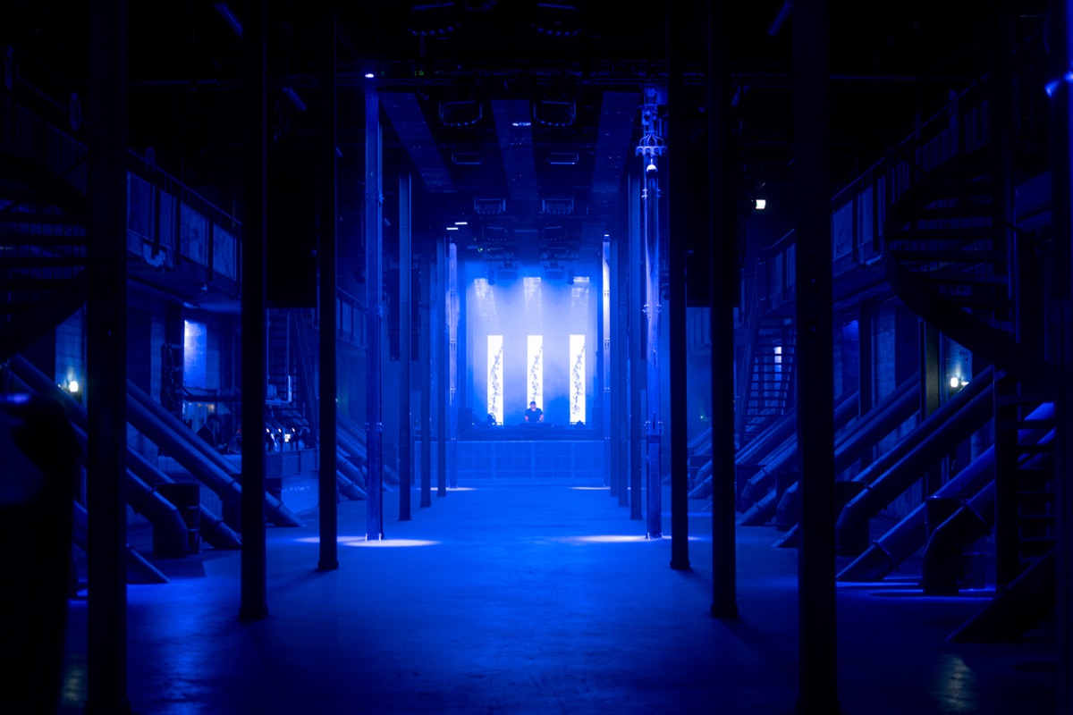 een betonnen hal in diepblauw licht, twaalf led-panelen rond een dj achterin